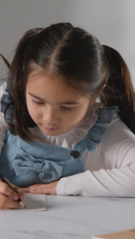 Vertical-Video-Studio-Shot-Of-Young-Girl-At-Table-Concentrating-On-Writing-In-School-Book-